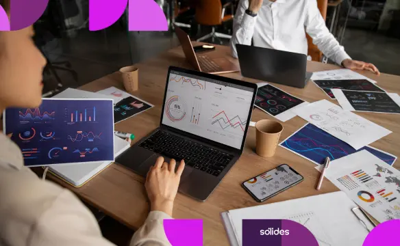 Analista de RH visualizando dashboards de indicadores em um laptop, cercada por relatórios e gráficos impressos sobre uma mesa de madeira, em um ambiente de escritório moderno. A imagem apresenta elementos gráficos em tons de roxo e rosa nas extremidades e o logotipo da Sólides no canto inferior direito.