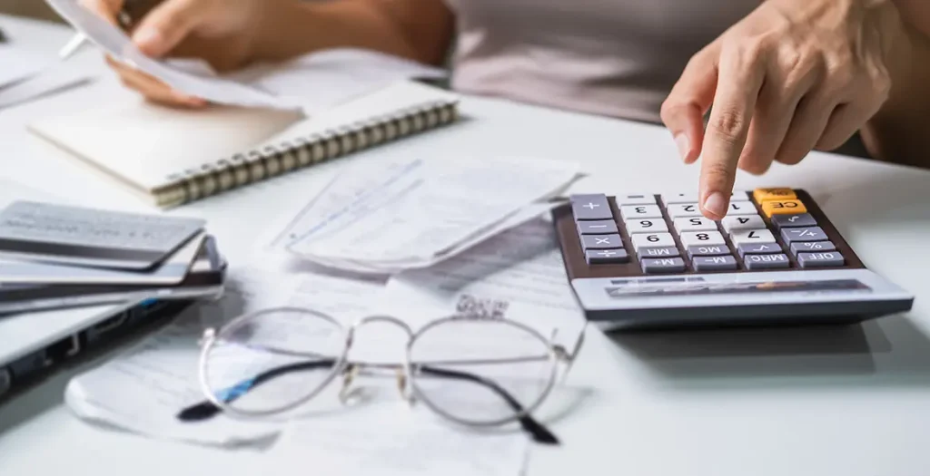 Mulher sentada a uma mesa utilizando uma calculadora com a mão direita e segurando um caneta com a mão esquerda, que também segura um papel ou recibo. Há óculos de leitura e outros papéis ou recibos espalhados na mesa, sugerindo que a pessoa está trabalhando com finanças ou fazendo um orçamento. A cena foca principalmente nas mãos da pessoa e nos itens sobre a mesa, sem mostrar o rosto ou a parte superior do corpo.
Simulando cálculo de salário