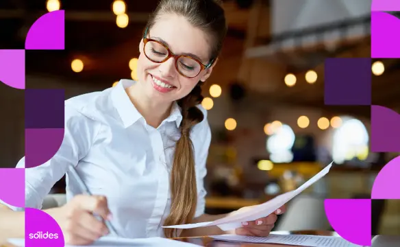 Mulher sorridente com óculos analisando relatórios manuais, representando a importância de monitorar os KPIs de Folha de Pagamento para garantir a saúde financeira da empresa com o Software de RH da Sólides.