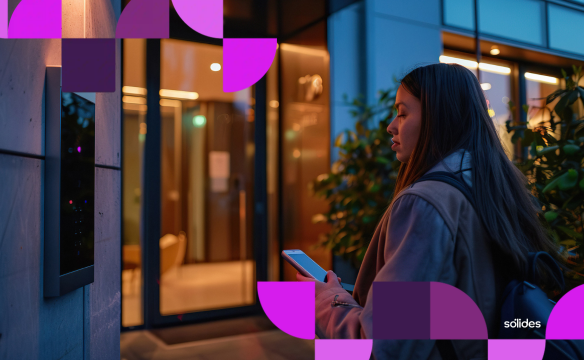 Mulher utilizando smartphone para registro de ponto com geolocalização em frente a um prédio comercial ao anoitecer. Logo da Sólides no canto inferior.