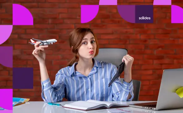 Colaboradora sorridente sentada em sua mesa de escritório na Sólides, segurando uma miniatura de avião em uma mão e um tablet na outra, simbolizando o retorno das férias ao trabalho com um notebook aberto à sua frente.
