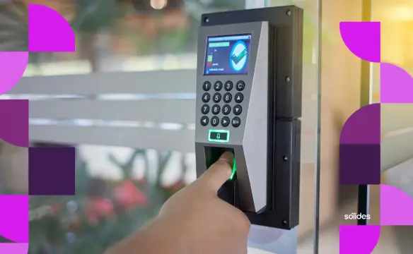Pessoa registrando o ponto através de um relógio biométrico com leitor de impressão digital em um ambiente de escritório. O equipamento possui teclado numérico e tela digital. Nas laterais da imagem, há elementos gráficos abstratos nas cores roxo e rosa, com o logotipo da Sólides no canto inferior direito.