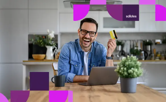 Homem sorridente de óculos e camisa jeans, sentado à mesa de uma cozinha moderna em frente ao notebook, segurando um cartão de benefícios corporativos, representando as regras de vale-alimentação e vale-transporte nas férias.