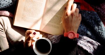 mão feminina segurando uma caneca de café e com um livro aberto em sua frente