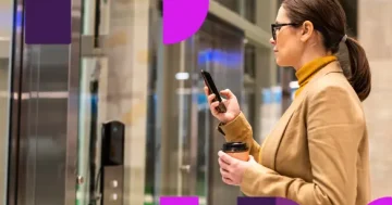 Mulher profissional de perfil, usando óculos e segurando um café, manuseia o celular para realizar o registro em um sistema de banco de horas ou conferir um acordo de compensação. A imagem possui grafismos abstratos em tons de roxo e rosa, com o logotipo da Sólides posicionado no canto inferior direito.