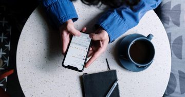 Mãos de uma pessoa segurando celular com rede social aberta sobre uma mesa em que há xícara, agenda e caneta.