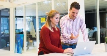 um homem e uma mulher, ela sentada e ele em pé ao seu lado, os dois olhando para o notebook