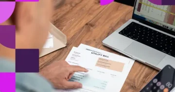 Visão superior de uma pessoa segurando um documento impresso de remessa sobre uma mesa de madeira, com notebook, calculadora e smartphone ao redor, realizando uma auditoria de folha de pagamento