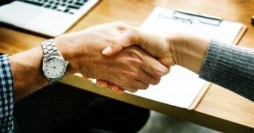 duas pessoas apertando as mãos, no fundo uma mesa com notebook e um documento sobre a mesa