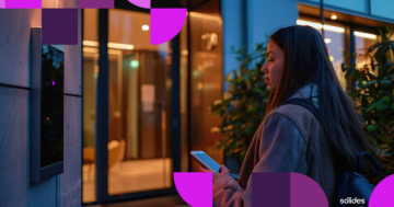 Mulher utilizando smartphone para registro de ponto com geolocalização em frente a um prédio comercial ao anoitecer. Logo da Sólides no canto inferior.