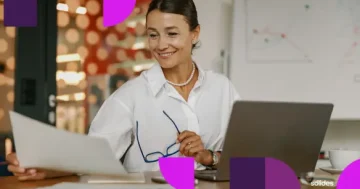 Mulher sorridente em escritório usando notebook e analisando documentos, representando a gestão de controle de ponto no trabalho híbrido.
