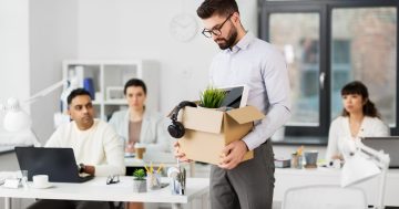 pessoas sentadas em suas mesas no escritório e um homem saindo com uma caixa com os seus pertences representando uma situação da demissão em massa
