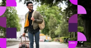 Homem negro sorridente, usando óculos, jaqueta verde e mochila, caminha ao ar livre em uma calçada arborizada puxando uma mala de viagem de rodinhas.