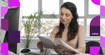 Uma mulher de cabelos escuros e compridos está sentada em sua mesa de escritório, concentrada na leitura de um documento em papel que segura com as duas mãos. À sua frente, há um computador desktop, um teclado, um mouse e um pequeno vaso com planta, idealizando como é importante realizar a integralização da folha de pagamento