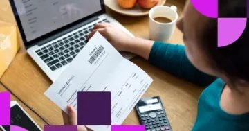 Visão superior de uma pessoa segurando um documento impresso de remessa sobre uma mesa de madeira, com notebook, calculadora e smartphone ao redor.