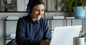 mulher sorrindo em frente ao computador, esta mulher utiliza um fone de ouvido azul e o computador é branco