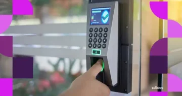 Pessoa registrando o ponto através de um relógio biométrico com leitor de impressão digital em um ambiente de escritório. O equipamento possui teclado numérico e tela digital. Nas laterais da imagem, há elementos gráficos abstratos nas cores roxo e rosa, com o logotipo da Sólides no canto inferior direito.
