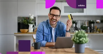 Homem sorridente de óculos e camisa jeans, sentado à mesa de uma cozinha moderna em frente ao notebook, segurando um cartão de benefícios corporativos, representando as regras de vale-alimentação e vale-transporte nas férias.