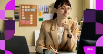 Mulher jovem, analista de Departamento Pessoal, usando óculos e blazer bege, trabalha concentrada em um escritório moderno com computador e mesa digitalizadora.
